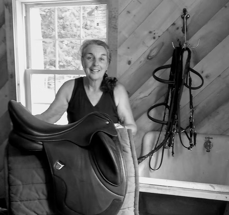 Woman in tack room smiles, holding saddle. Bridle hangs nearby. Window in background.