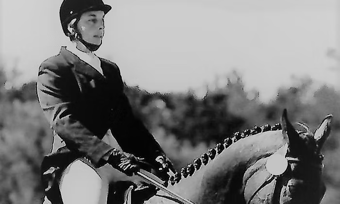 Woman on horseback in riding attire, outdoors. Horse has braided mane; woman holds reins.