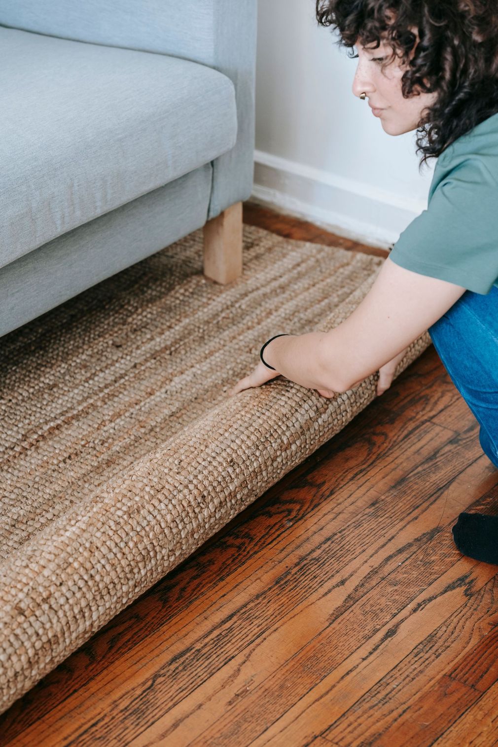 Person rolling up a woven rug beside a gray couch on a hardwood floor