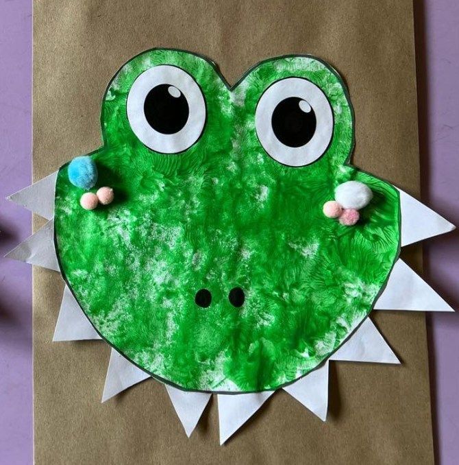 Green alligator craft with white teeth, large eyes, and pompom details, on a brown paper bag.