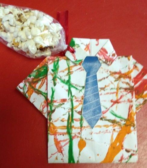 A colorful, origami shirt with a blue tie next to a bag of popcorn on a red surface.