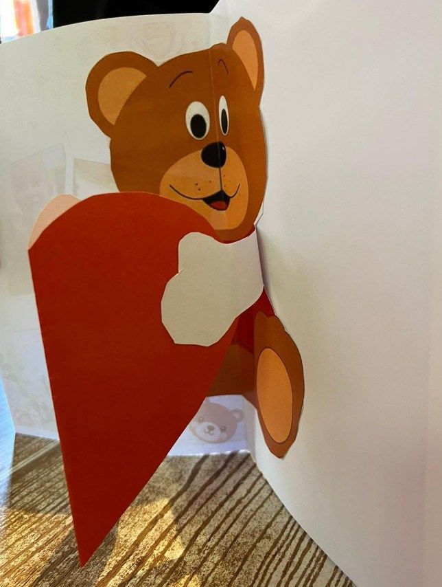 Brown teddy bear pop-up card holding a red heart. White background.