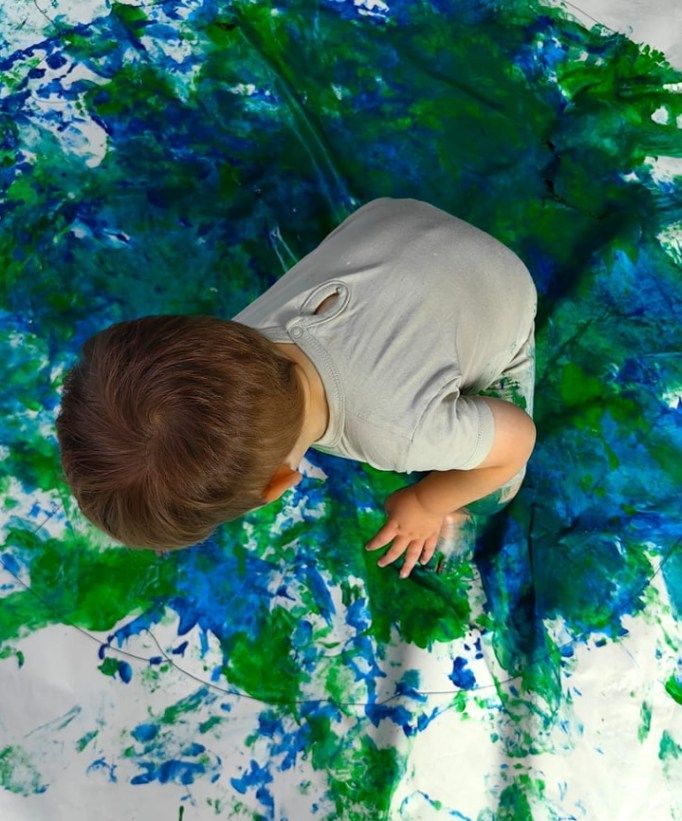 Child painting with blue and green paint on a white sheet, seen from above.