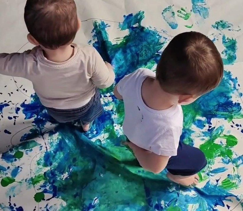 Two young children painting on large white paper with blue and green paint.