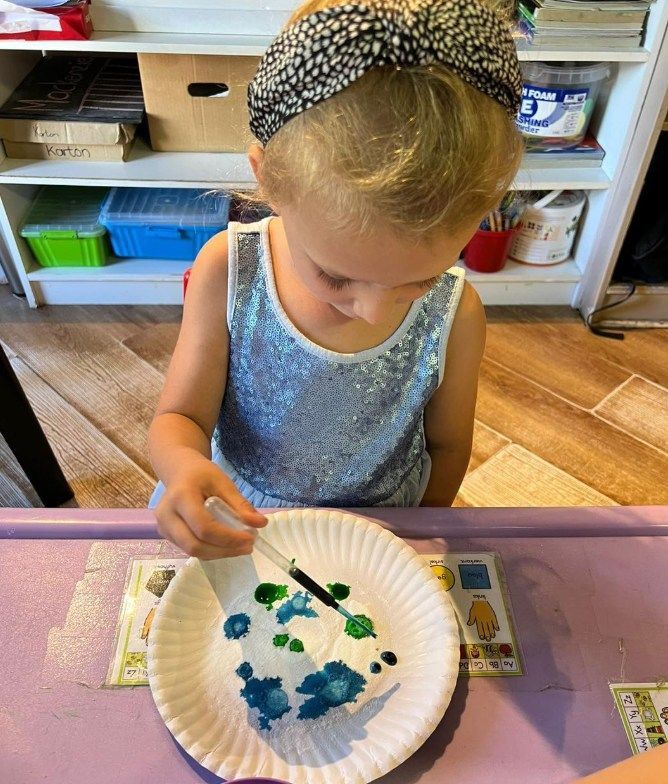 Girl with headband, using a dropper, drops green and blue liquid onto a white plate on a purple table.