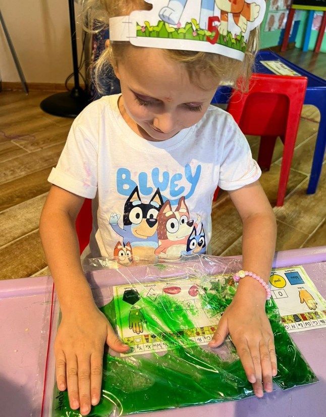 Girl in Bluey shirt wearing a birthday crown, playing with a green sensory bag at a table.
