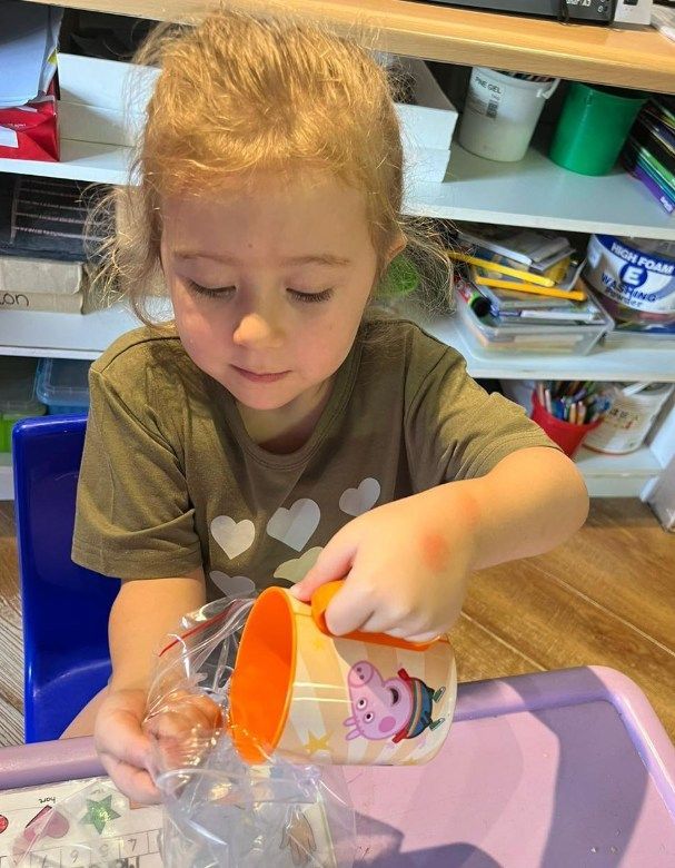 Girl pouring contents from Peppa Pig cup into a plastic bag. She is sitting at a table with shelves in the background.