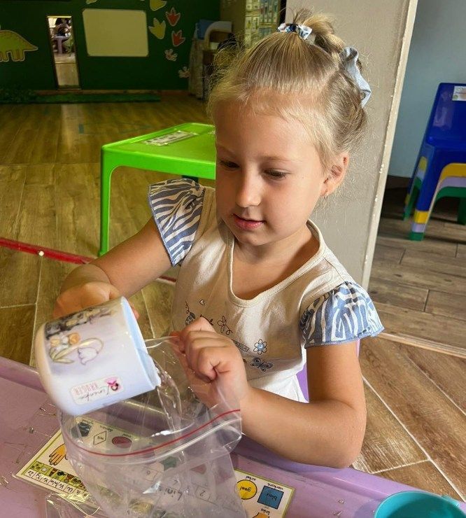 Girl pouring from a container into a plastic bag, in a brightly colored classroom.