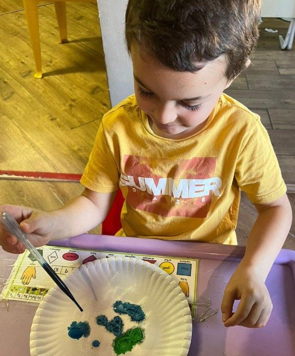 Boy in yellow shirt using a dropper to add liquid to a paper plate with colorful spots, indoors.