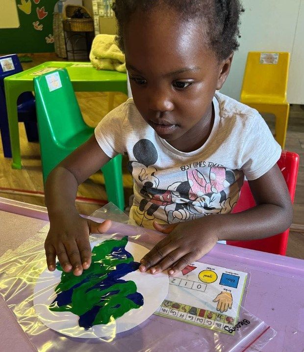 Young child focused on applying paint to a paper plate. Indoors at a table, green, blue paint.