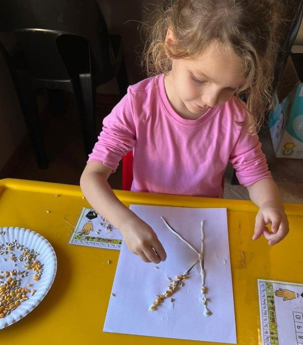 Young child in pink shirt doing craft project with corn kernels at a table.