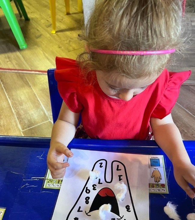 A young child in red shirt is playing with a worksheet and cotton balls at a table.