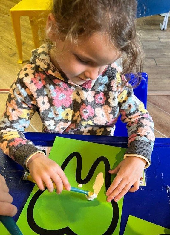 Girl painting a black outline on green paper with a toothbrush at a blue table.