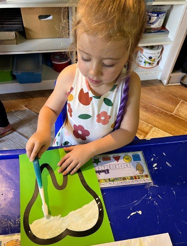 Young child painting white paint onto a green tooth cutout at a blue table.