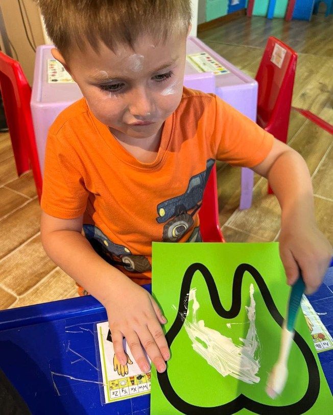 Boy painting a tooth on green paper with a toothbrush and white paint.