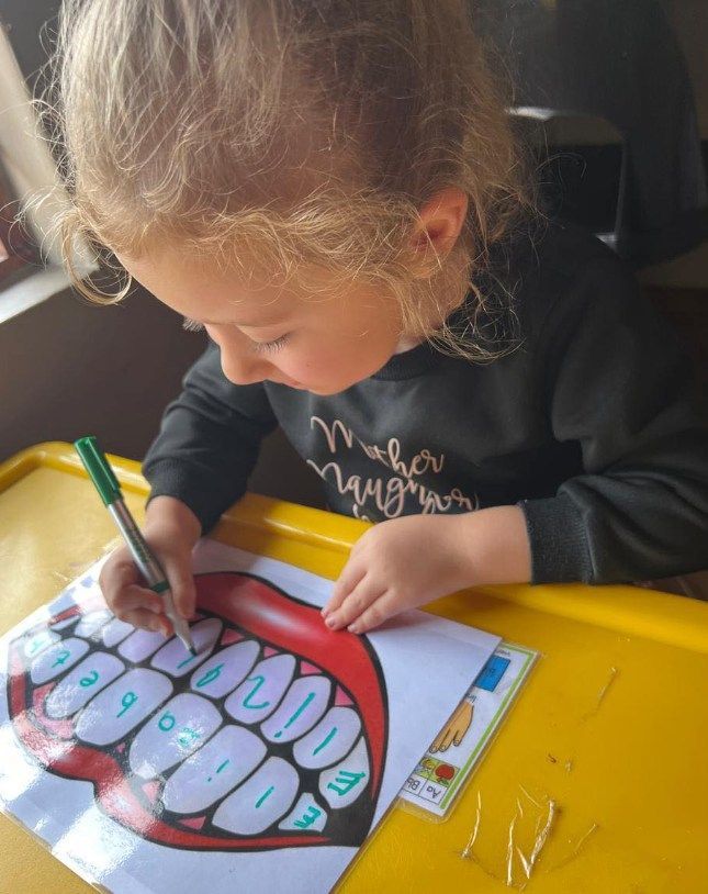 Child coloring teeth on a printed chart with a green marker, sitting at a yellow table.