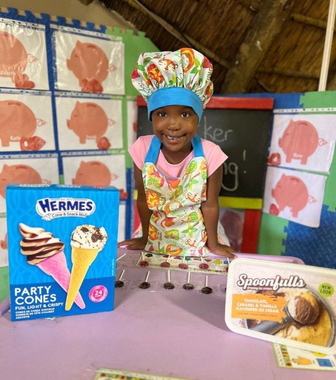 Young person in a chef's hat and apron smiles behind ice cream cones and toppings.