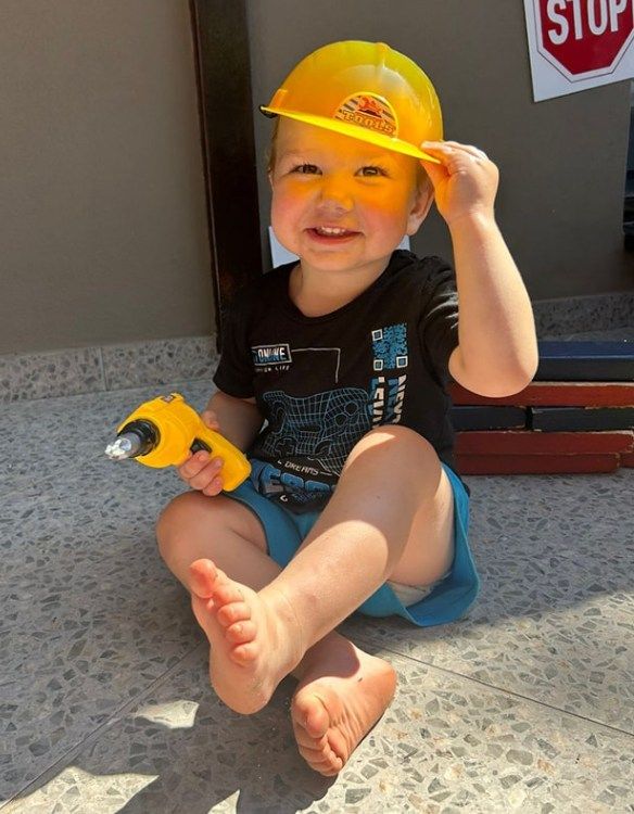 Young child in yellow hard hat holding a toy drill, smiling, sitting on a tiled surface.