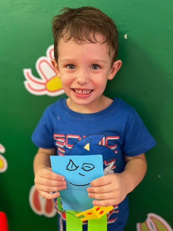 Boy holding a blue paper monster craft, smiling at the camera. Green background with decorations.