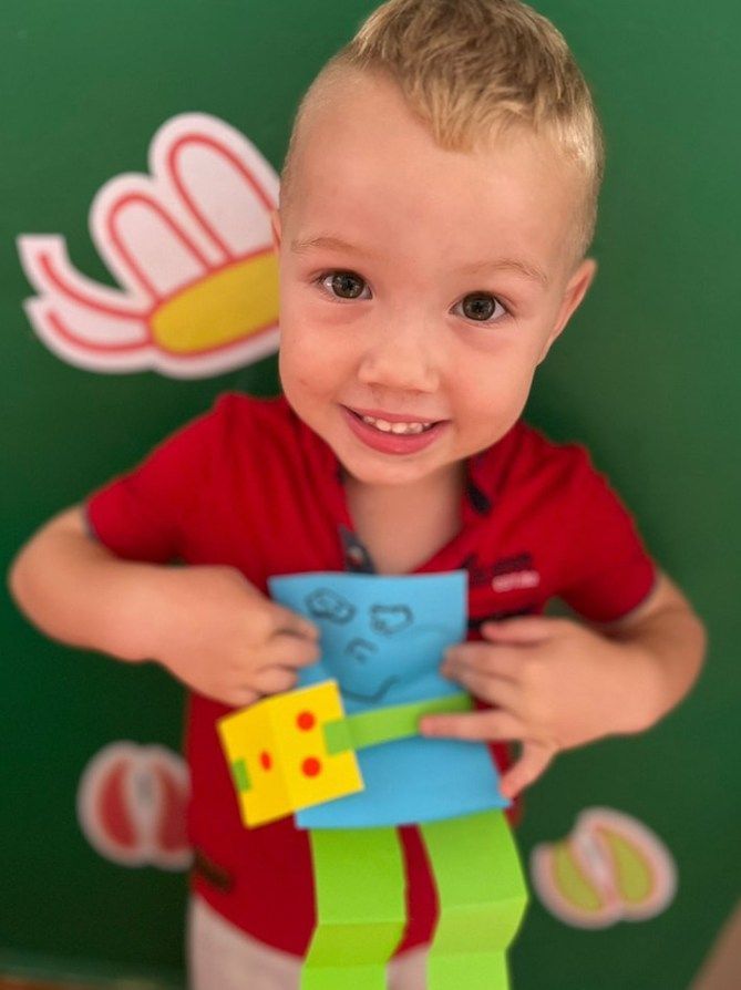 Child holding a blue paper craft zombie, smiling, in front of a green background with floral decorations.