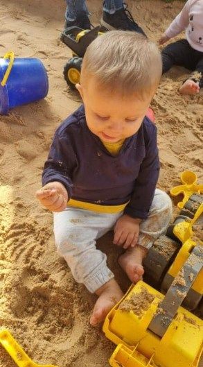 Child playing in a sandbox with yellow toy trucks. Wearing blue shirt and white pants, looking down.