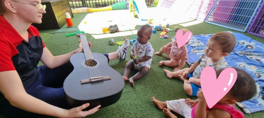 A woman sits with babies on a green surface, holding a black guitar. Children look at the guitar.