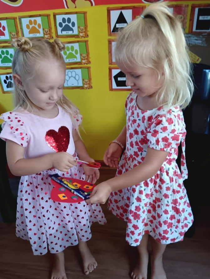Two young girls in heart-print dresses exchange a Valentine's card in front of a colorful wall.