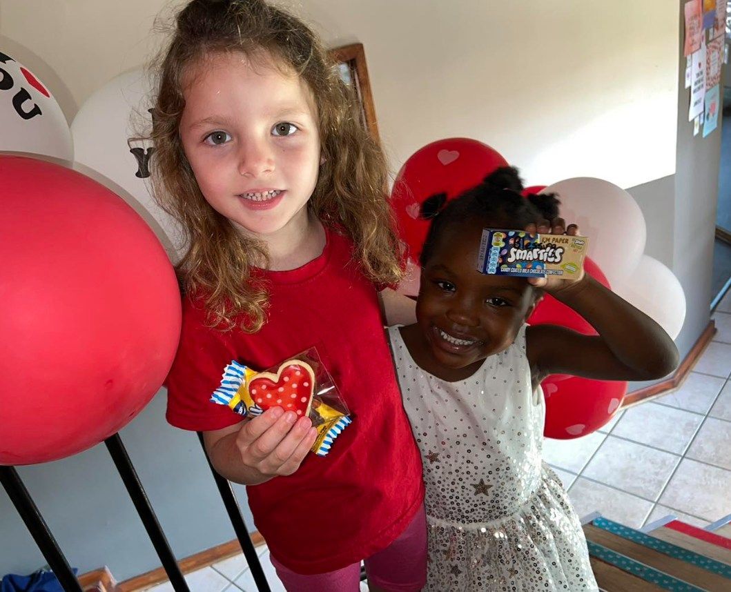 Two girls smiling, holding candy and a card; red and white balloons in the background.