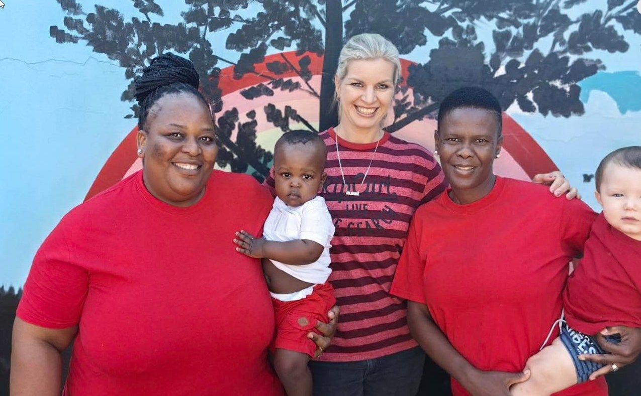 Five people stand in front of a mural. Two women hold babies, all wearing red. The other woman wears stripes.