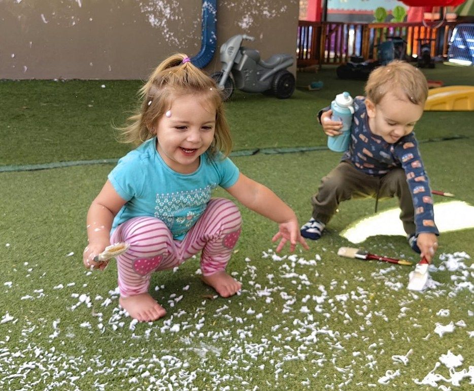 Two children playing in artificial snow on green turf, smiling.