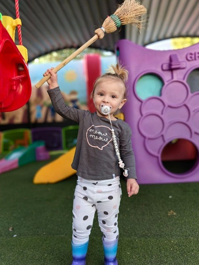 Young child holds a broom up. They wear a gray shirt, spotted pants, and boots, with a pacifier. Playground background.