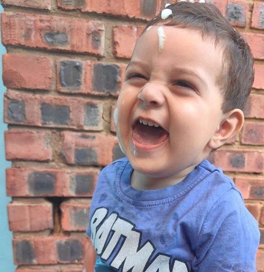 A toddler with frosting on his hair and face laughs in front of a brick wall.