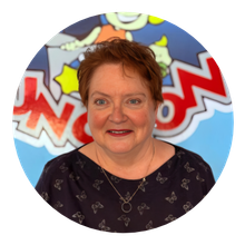 Woman with reddish-brown hair smiles, wearing a pink floral shirt, beaded necklace; background with papers.
