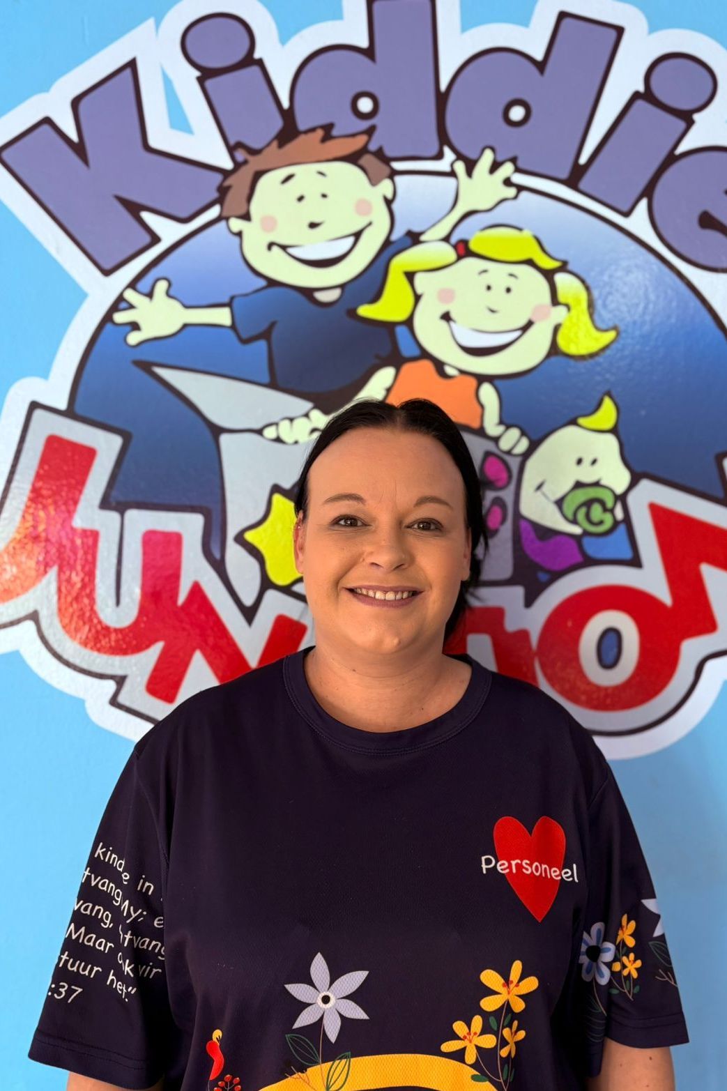 Woman in blue shirt smiles in front of a Kiddie World logo with cartoon children.