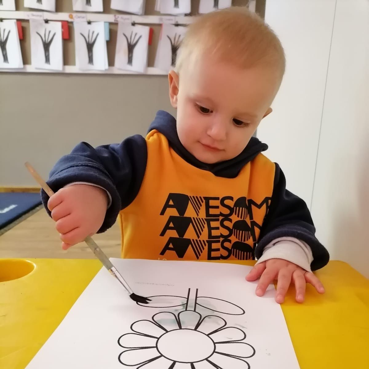 A young child painting a black daisy on white paper at a yellow table; inside a classroom.