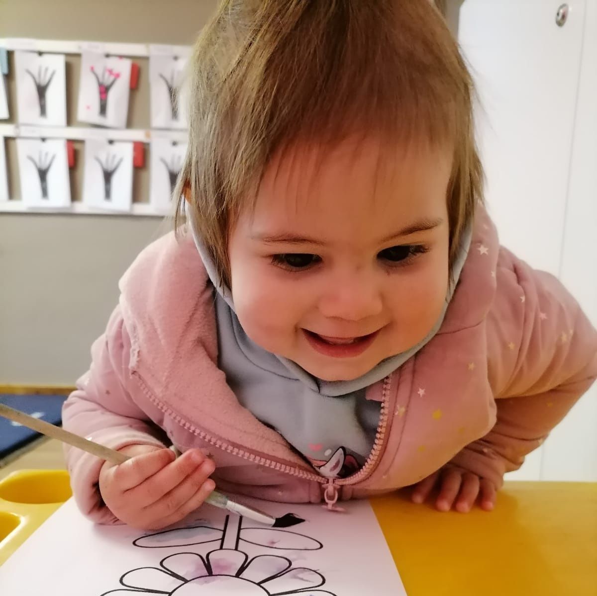 Smiling child in pink jacket painting a flower at a table.