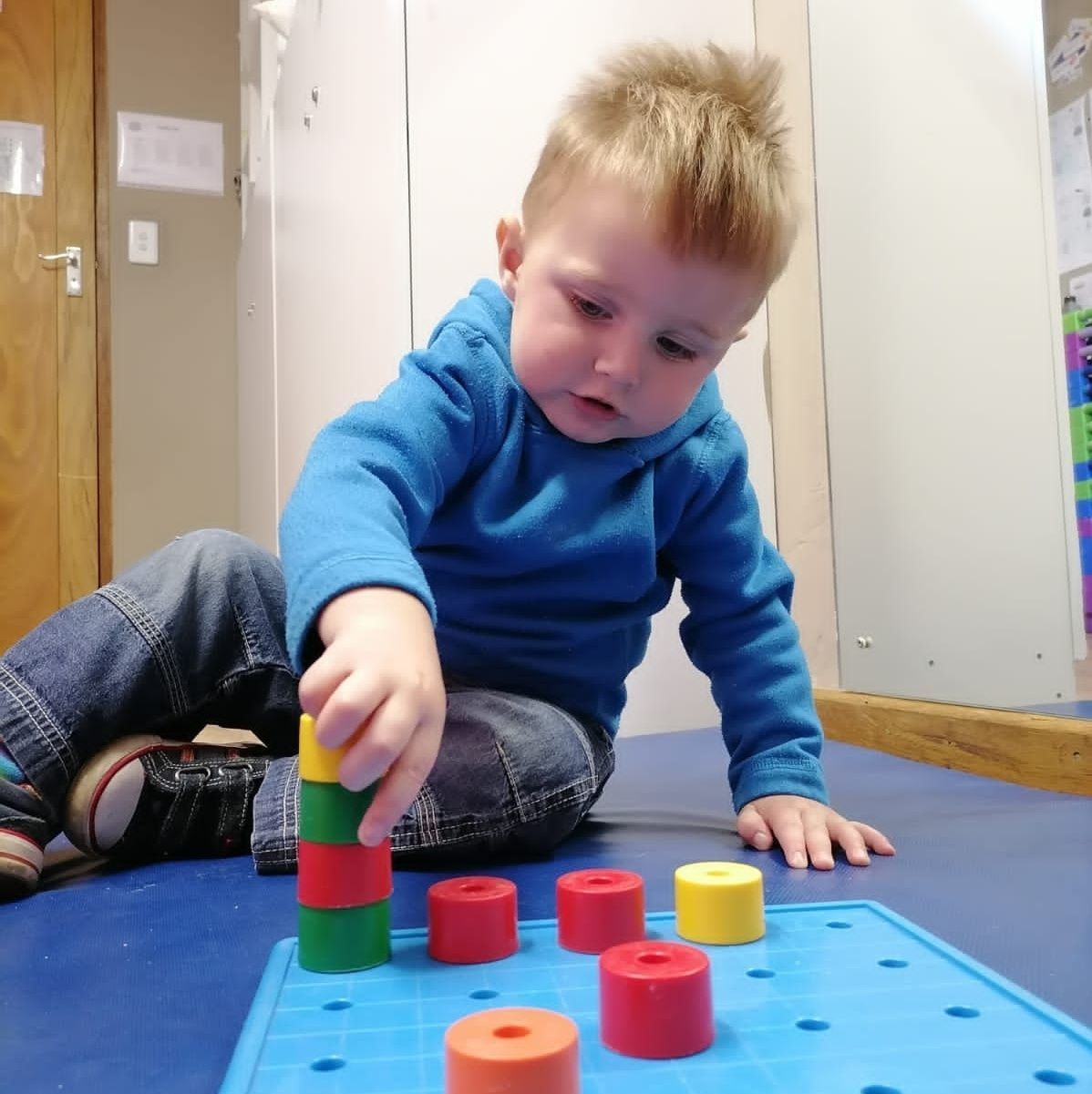 Young child stacking colorful blocks on a blue pegboard, indoors.