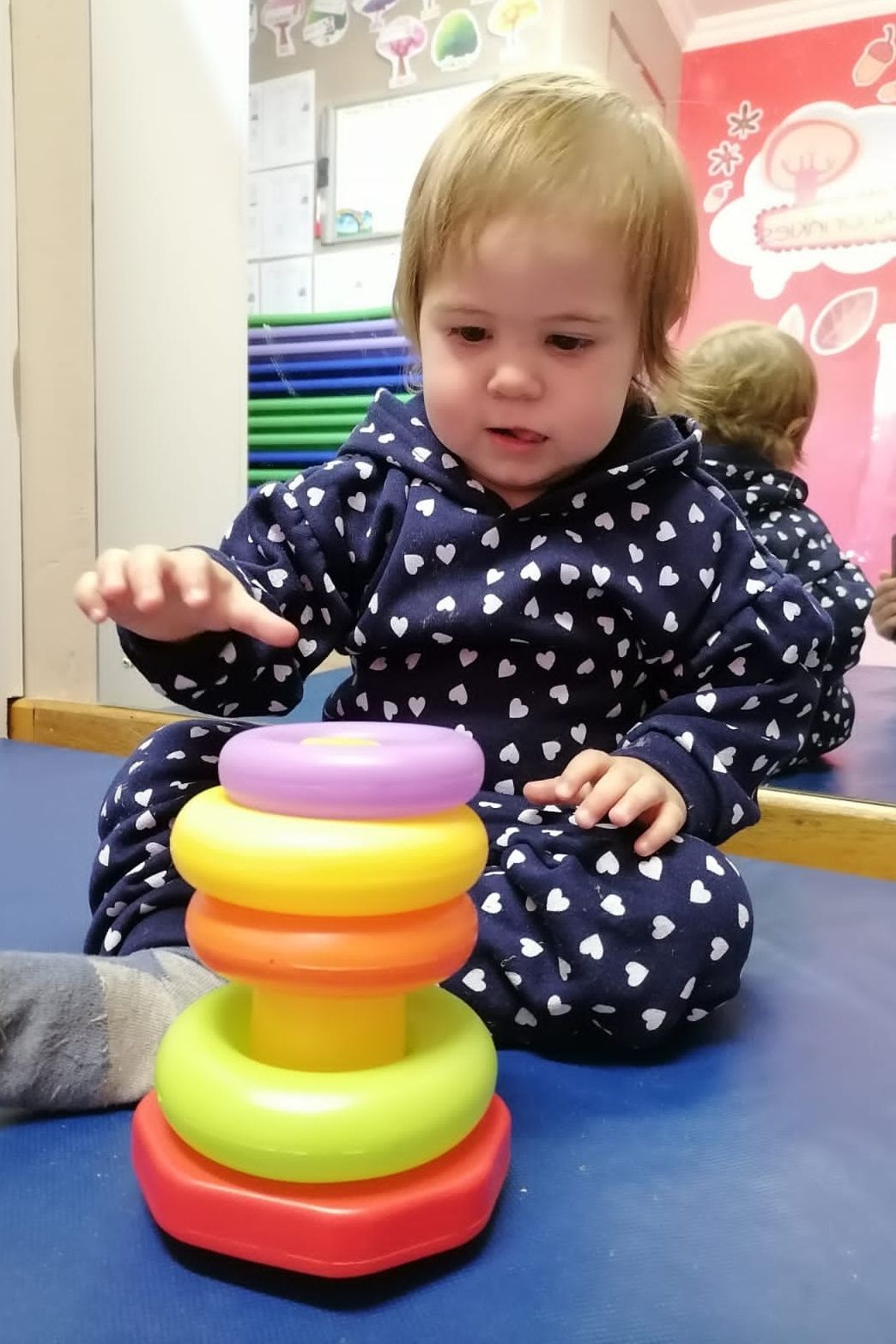 Child in blue heart-print outfit stacking colorful rings on a toy tower, with a mirror reflection.