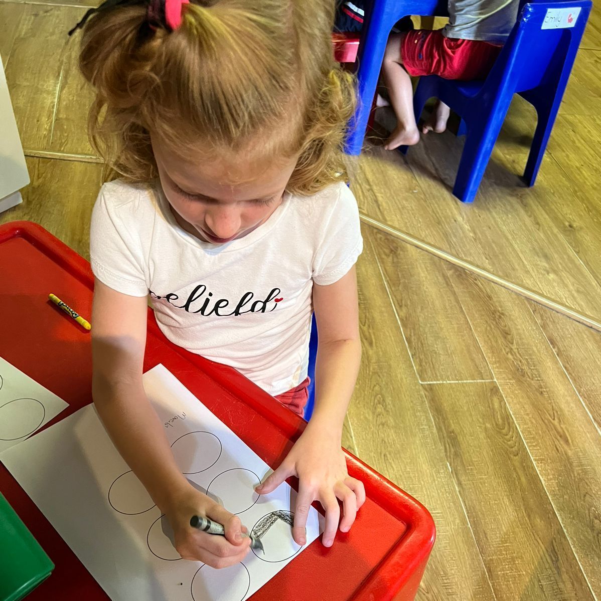 Girl seated at a red table, drawing inside a circle with a marker. Other child and blue chair visible.