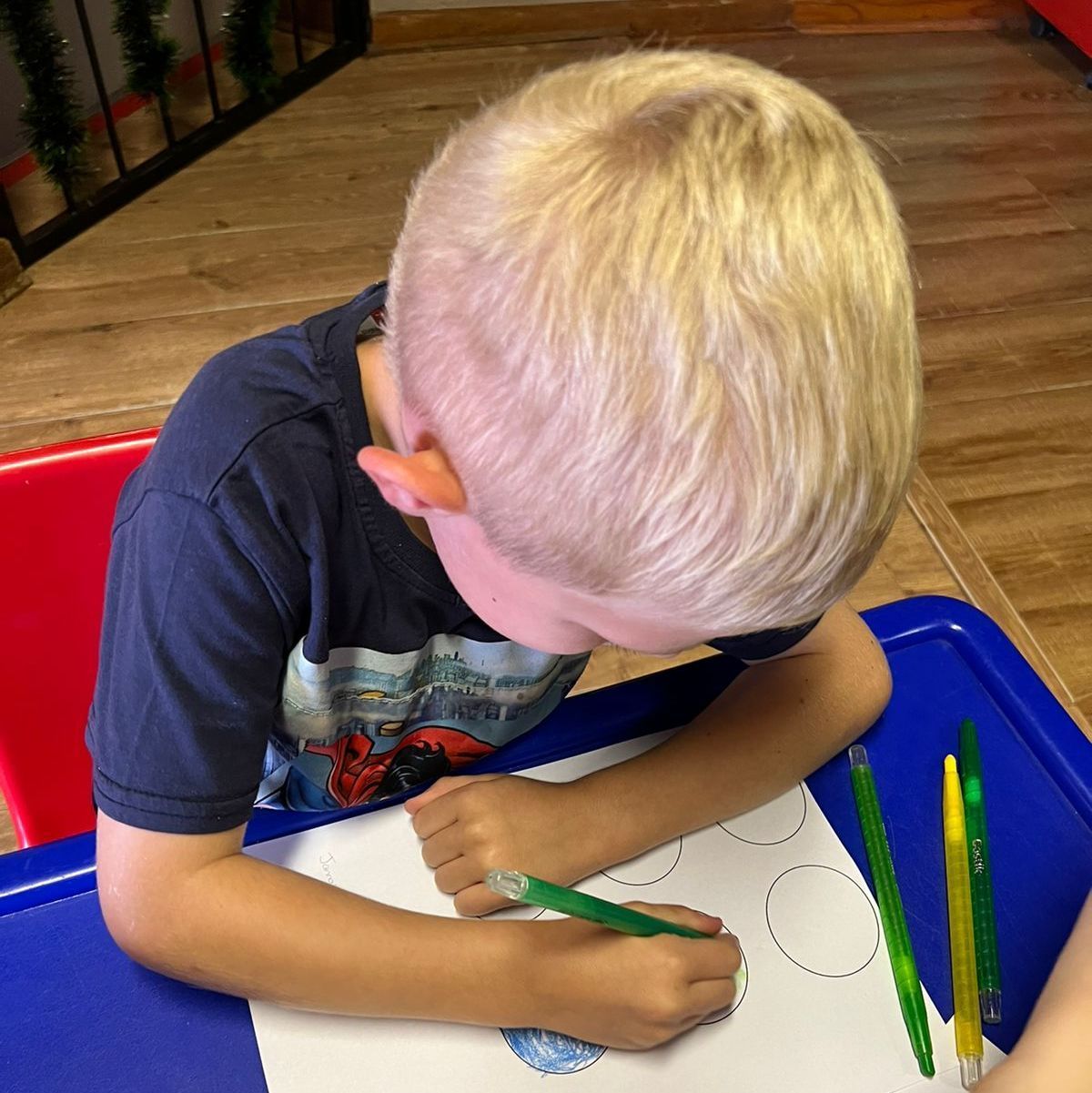 Blond-haired child seated at a blue table, coloring a drawing with green pencil.