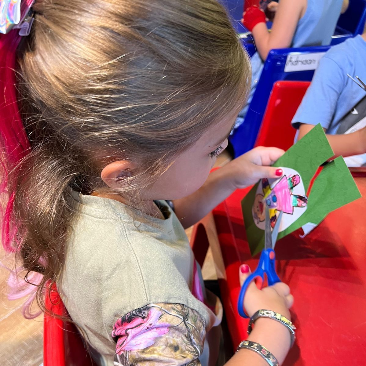 Young child with brown hair cuts green paper with blue scissors at a red table.