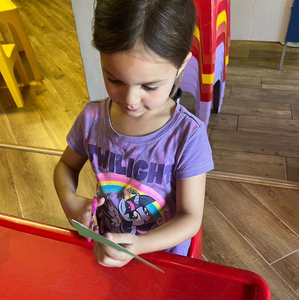 Young child in purple shirt cutting paper at a red table.