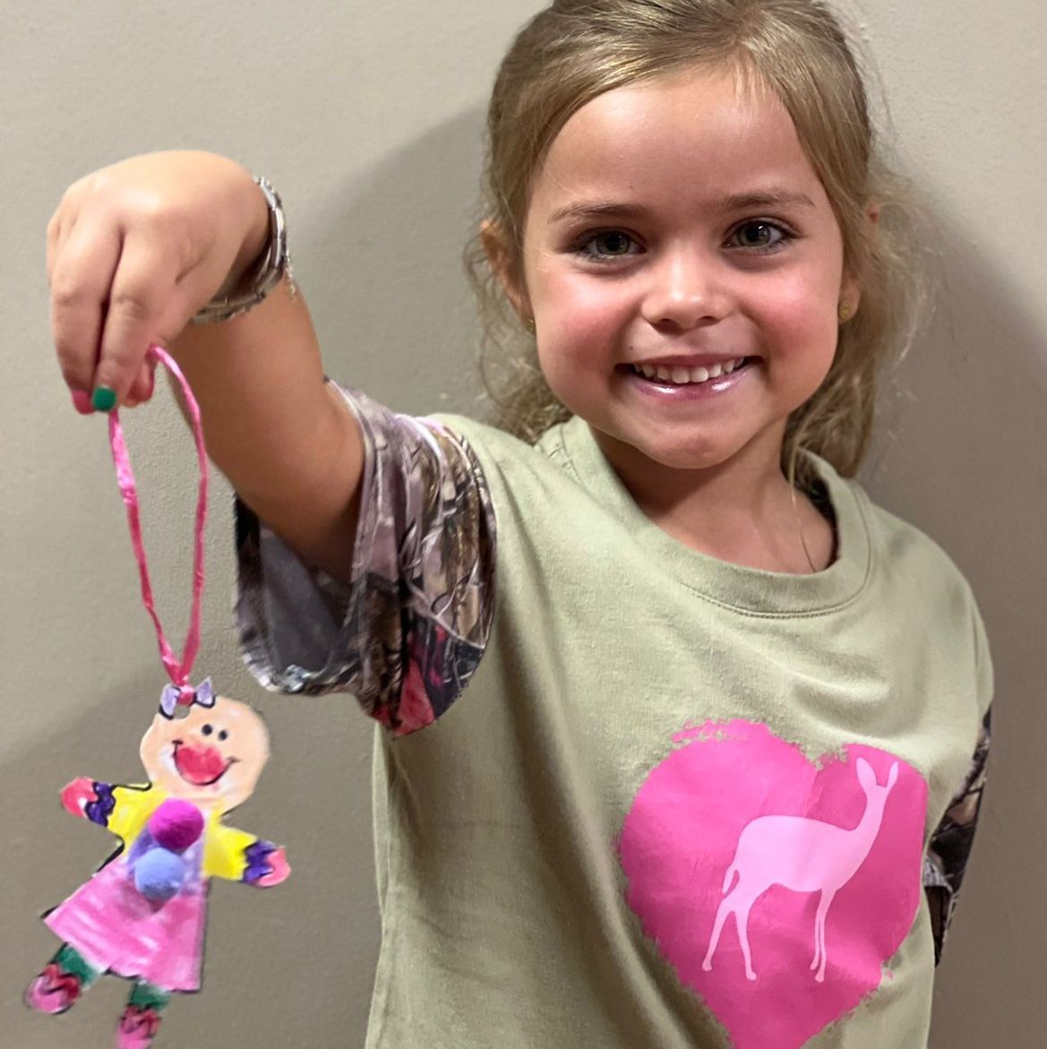 Girl smiles, holding up handmade doll necklace with pink heart shirt.