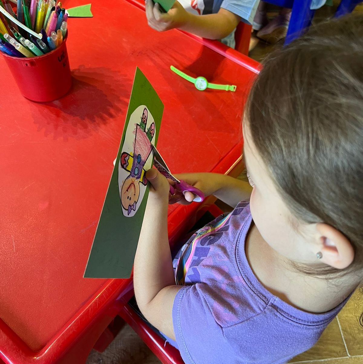Child cutting paper with scissors at a red table. There is a pencil holder and other supplies nearby.
