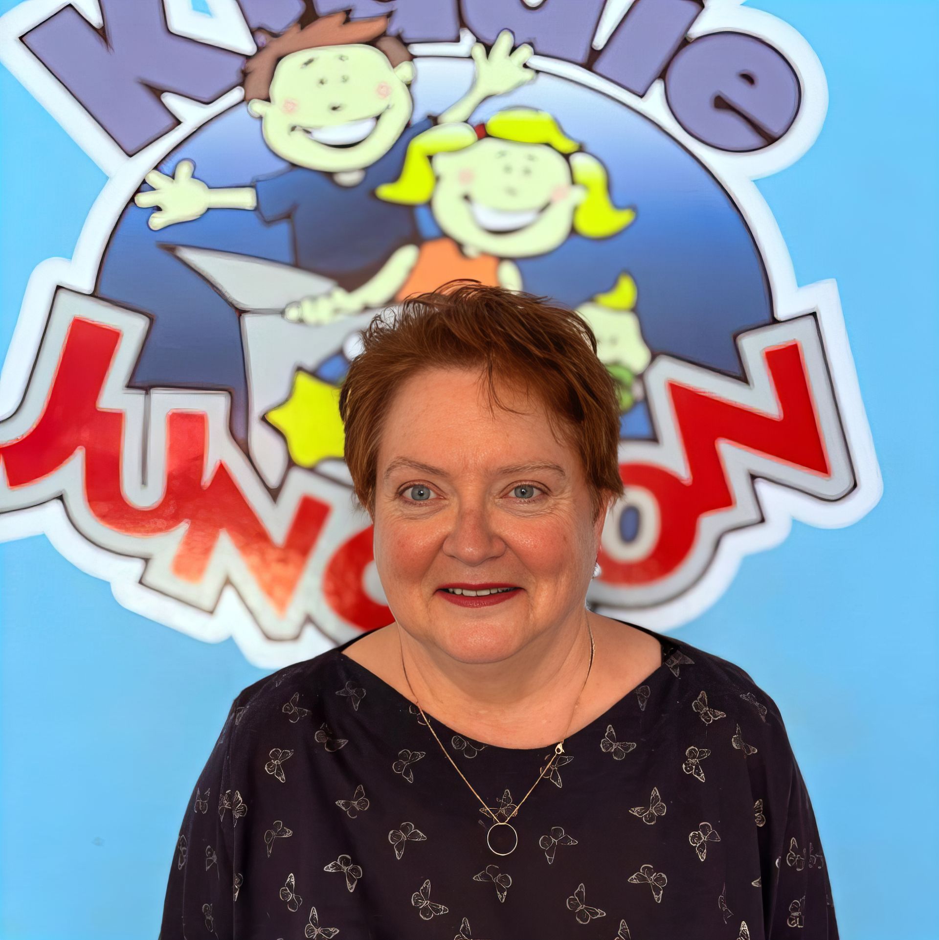 Woman with short red hair smiles, sits at desk with tablet, laptop, mug. Pink floral shirt. Bulletin board in background.