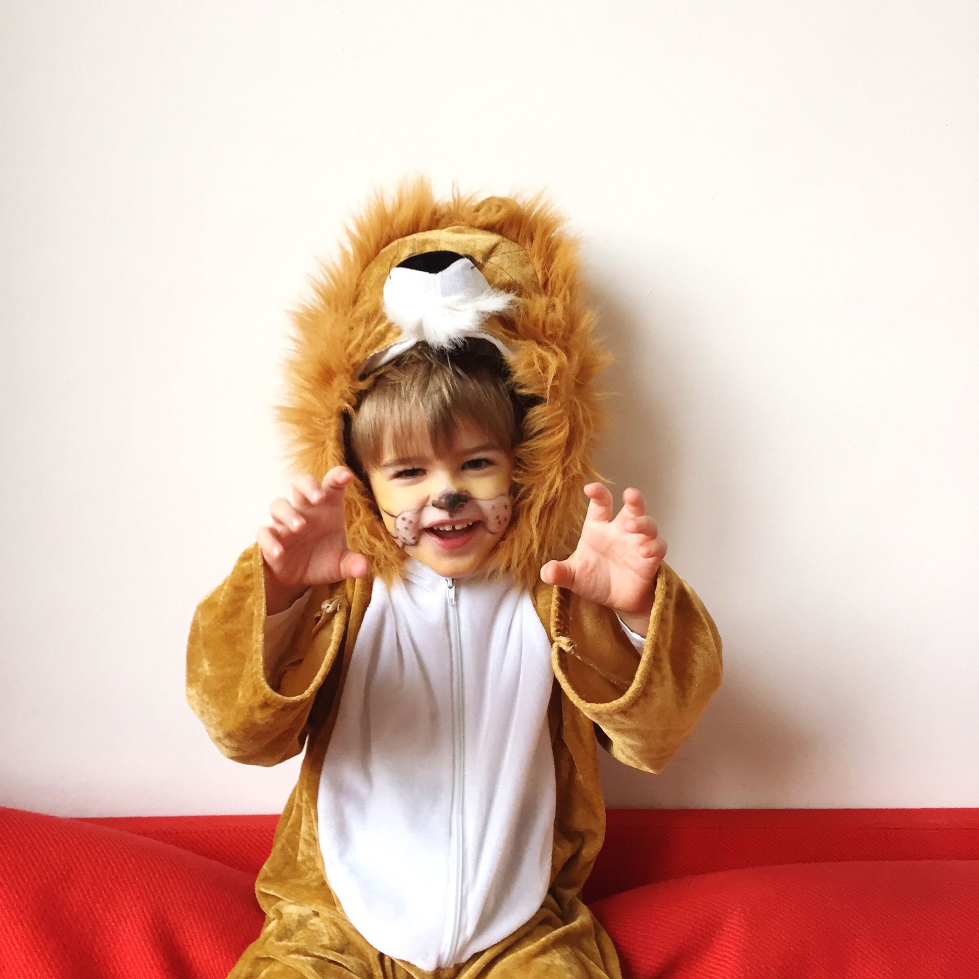 Child in lion costume smiles, raises hands, and sits on a red surface against a white wall.