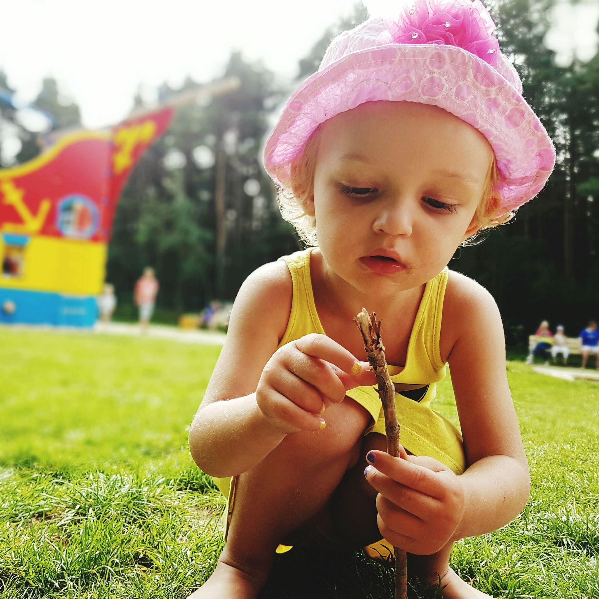Young child in pink hat and yellow dress examines a stick outdoors on a sunny day.