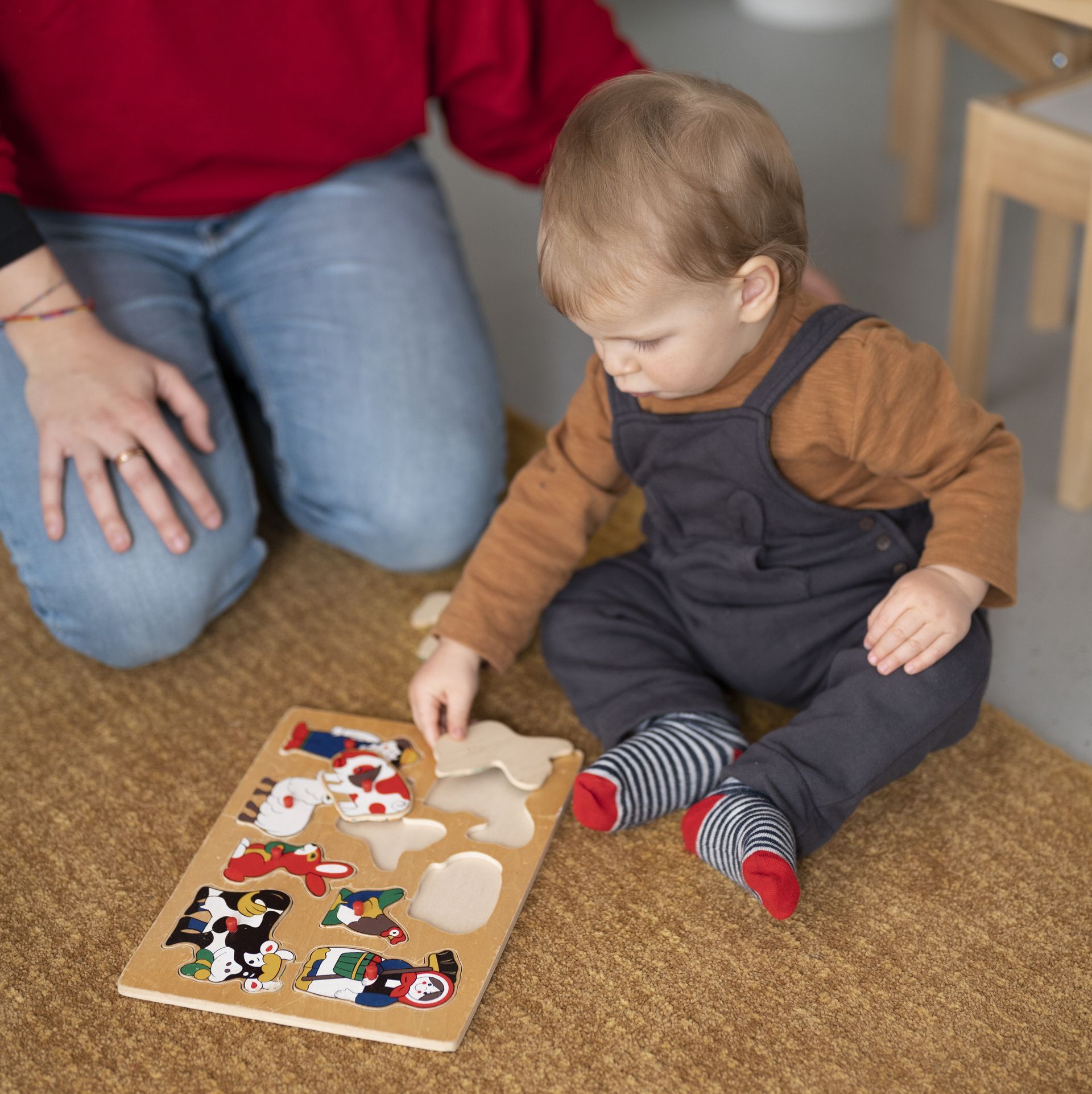 Child playing with a wooden puzzle of vehicles on a rug; adult watches nearby.