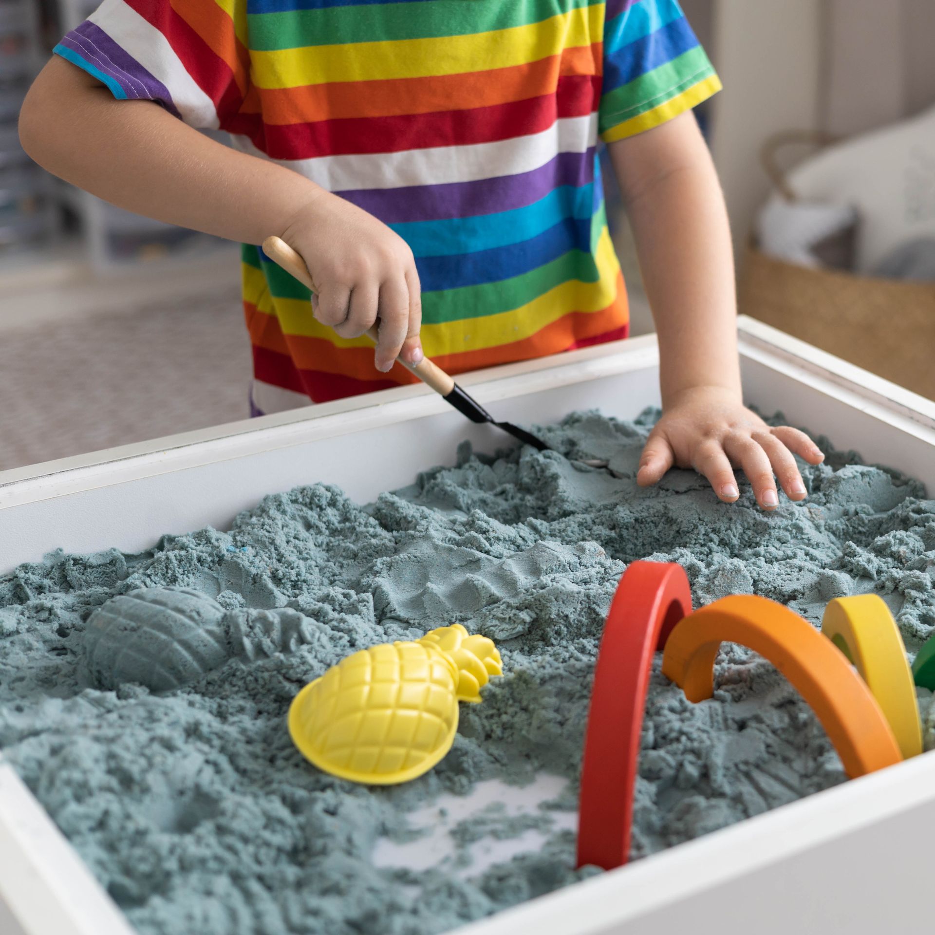 Child playing in a sandbox with blue-gray sand, using a small shovel. Includes toy pineapple and rainbow arch.
