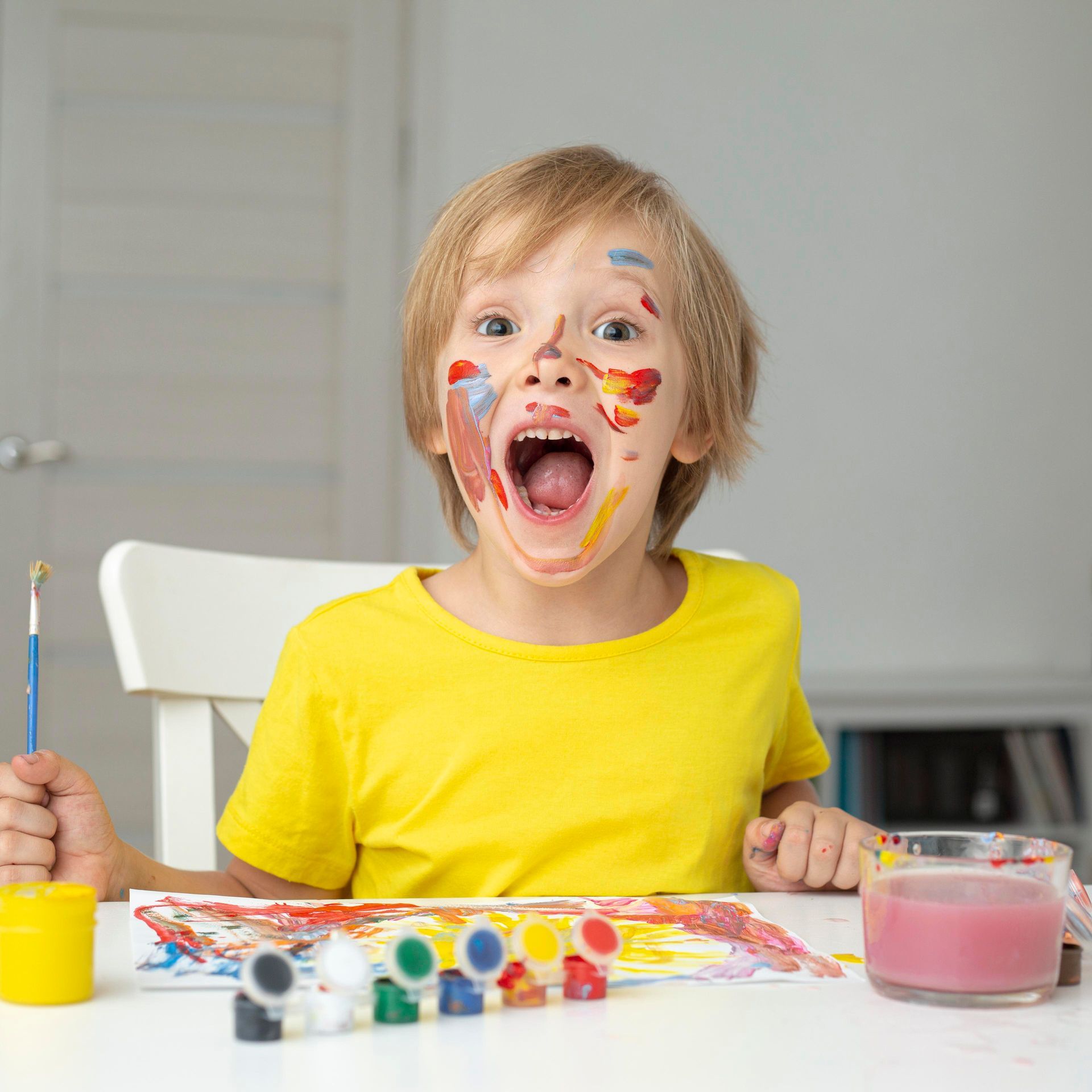 Child with paint on face, open mouth, holding brush, table with paints.